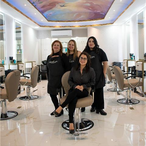 A group of four women in a salon setting, with two standing and two seated, all wearing matching aprons. The salon features elegant decor, including modern styling chairs and bright lighting.